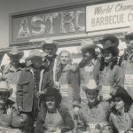 A group of people posing in front of a world's championship bar-b-que contest sign at the Houston Rodeo.
