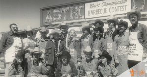 A group of people posing in front of a world's championship bar-b-que contest sign at the Houston Rodeo.
