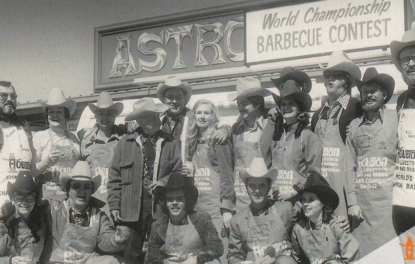 A group of people posing in front of a world's championship bar-b-que contest sign at the Houston Rodeo.